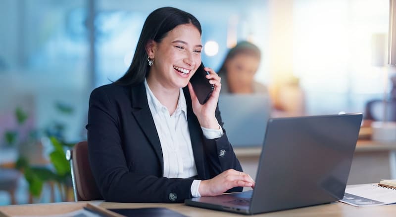 A businesswoman in professional attire smiling while taking a phone call at her desk with a laptop, representing modern internet phone service in a bright office setting.